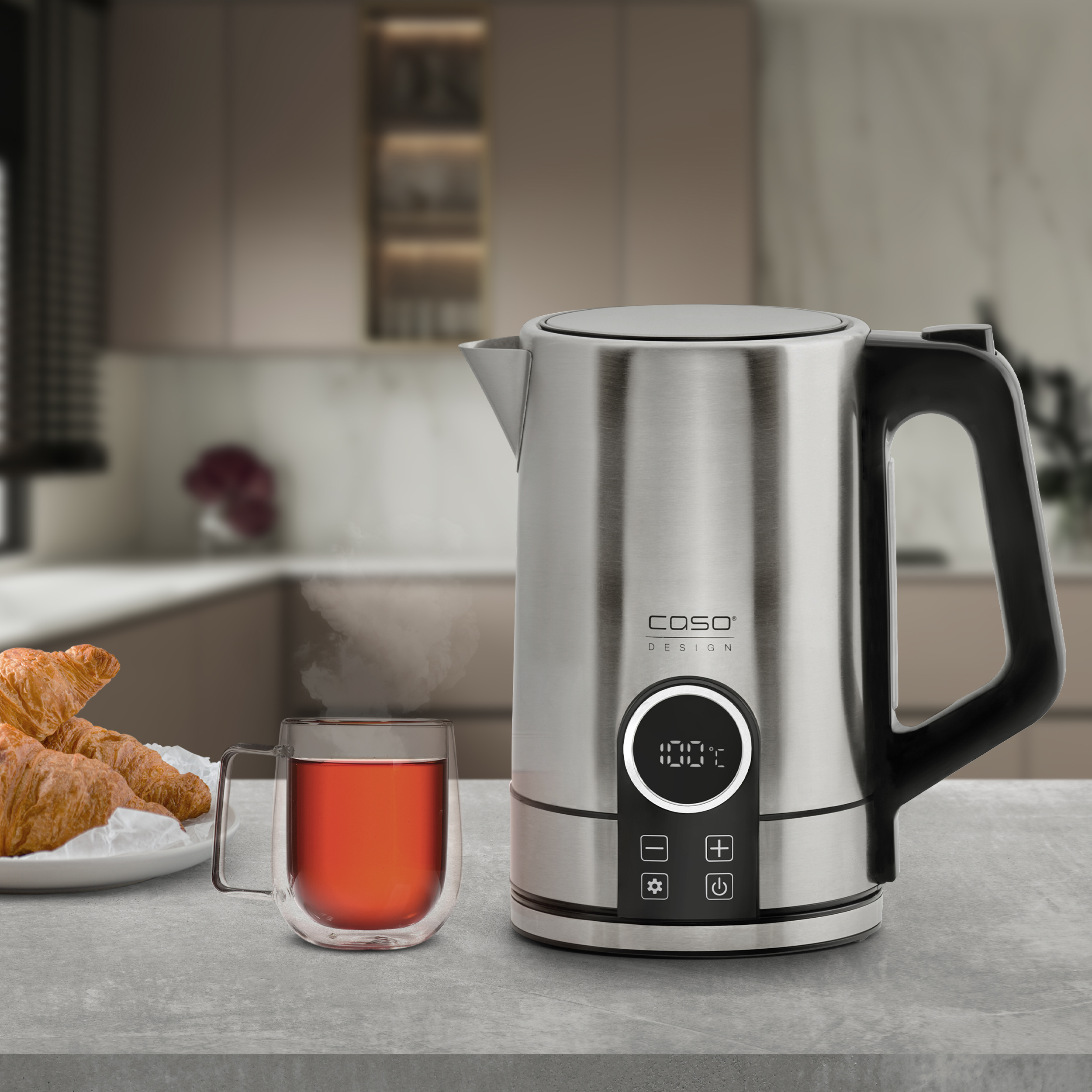 Un bollitore elettrico in acciaio inossidabile con manico nero, accanto a una tazza di tè e croissant.