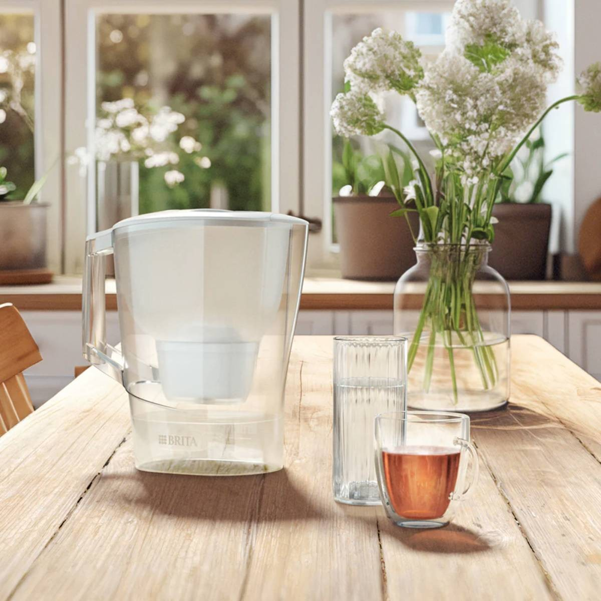 Auf einem Tisch stehen ein Wasserfilter, Gläser mit Wasser und Tee sowie eine Vase mit weißen Blumen. Im Hintergrund ist ein Fenster.