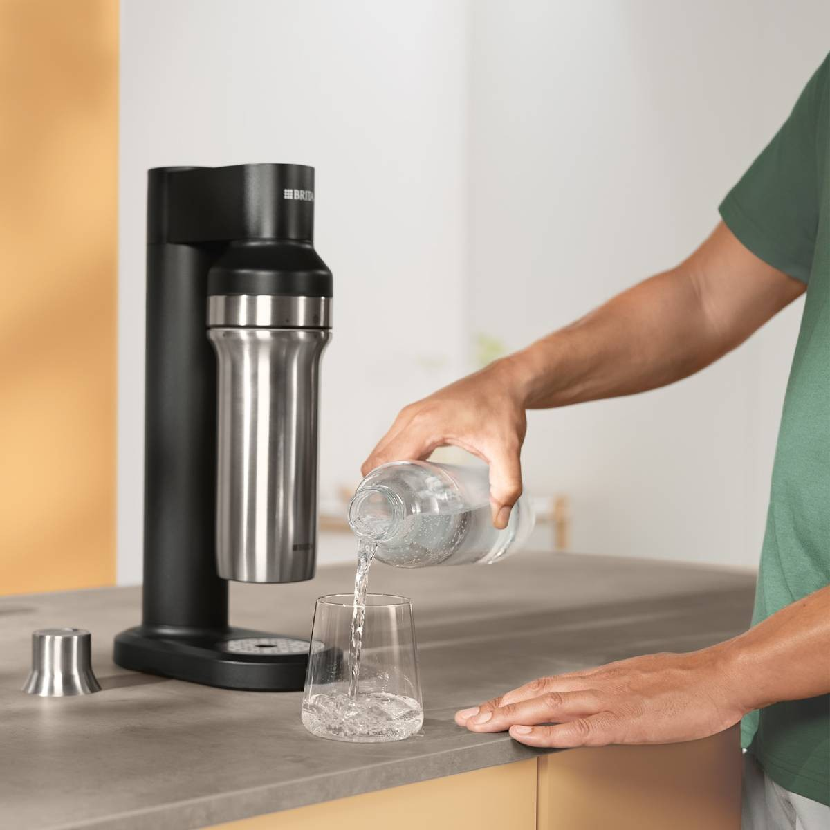 Eine Person gießt Wasser aus einer Flasche in ein Glas. Links steht ein schwarzer Wasserspender. Daneben ein kleiner Metallbecher.