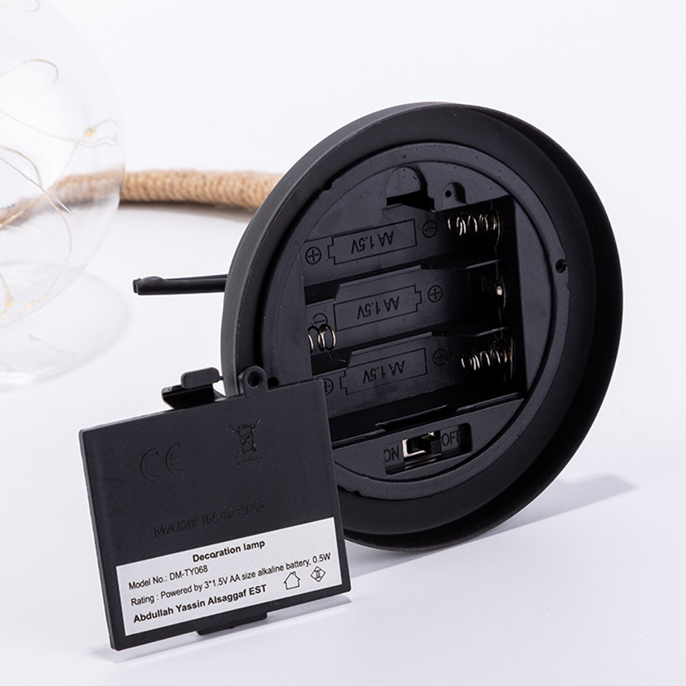 Schwarzer Lampensockel mit geöffnetem Batteriefach. Ein Batteriedeckel ist geöffnet. Eine Glaskugel mit Lichtern und Seil im Hintergrund.