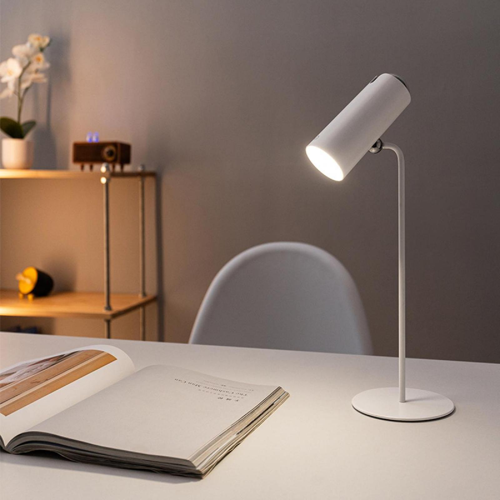 Eine weiße Schreibtischlampe beleuchtet ein offenes Buch. Ein Holzregal enthält Dekoration. Im Hintergrund ist ein weißer Stuhl zu sehen.