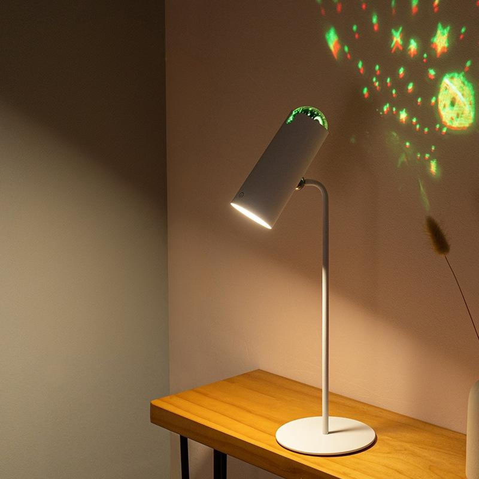 Eine weiße Lampe steht auf einem Holztisch und projiziert grüne und rote Lichter an die Wand. Eine Vase steht auf dem Tisch.