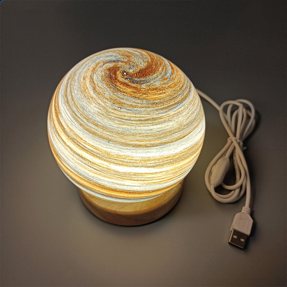 Eine dekorative Lampe in Form eines Planeten mit Holzsockel und USB-Kabel. Der Planet hat Wirbel aus Braun, Beige und Weiß.