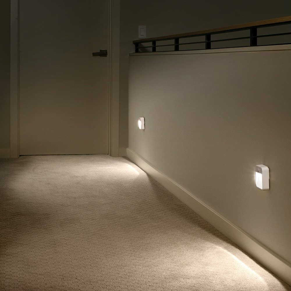 Ein Flur mit Teppich, Wandleuchten und einer Tür. Die Lichter beleuchten Wand und Boden. Im Hintergrund ist ein Geländer zu sehen.