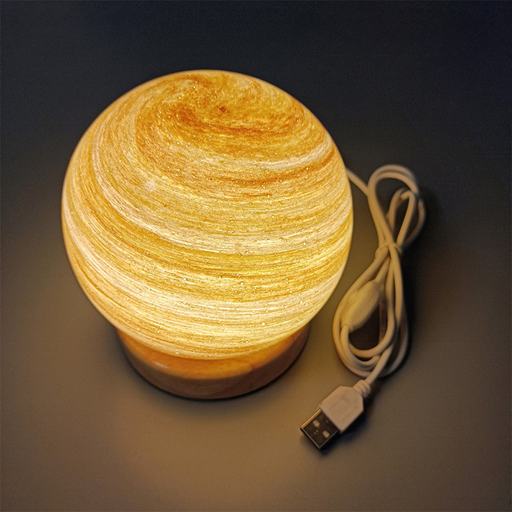 Eine kugelförmige Lampe mit einem Wirbelmuster aus Gelb und Braun. Sie steht auf einem Holzsockel und hat ein weißes USB-Kabel.