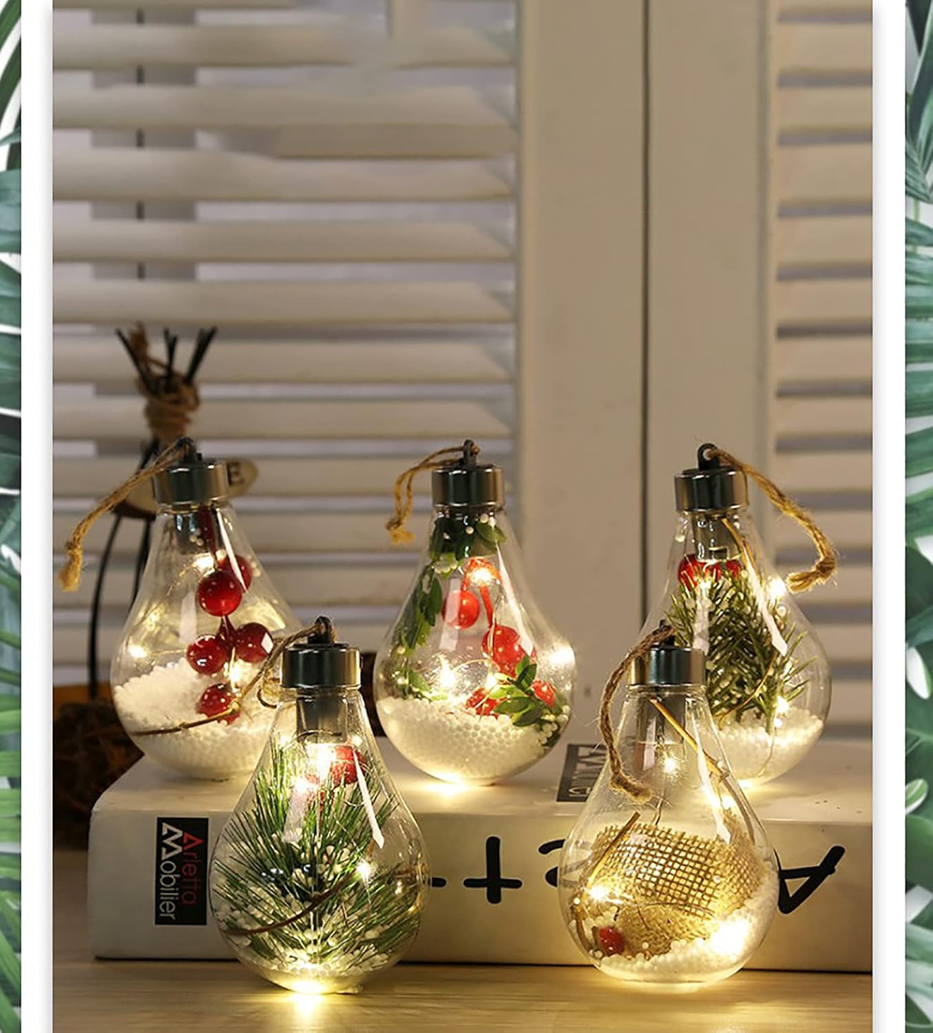 Fünf Glaszwiebeln mit Weihnachtsdekorationen und Lichtern. Sie stehen auf einer weißen Oberfläche mit verschwommenem Jalousienhintergrund.