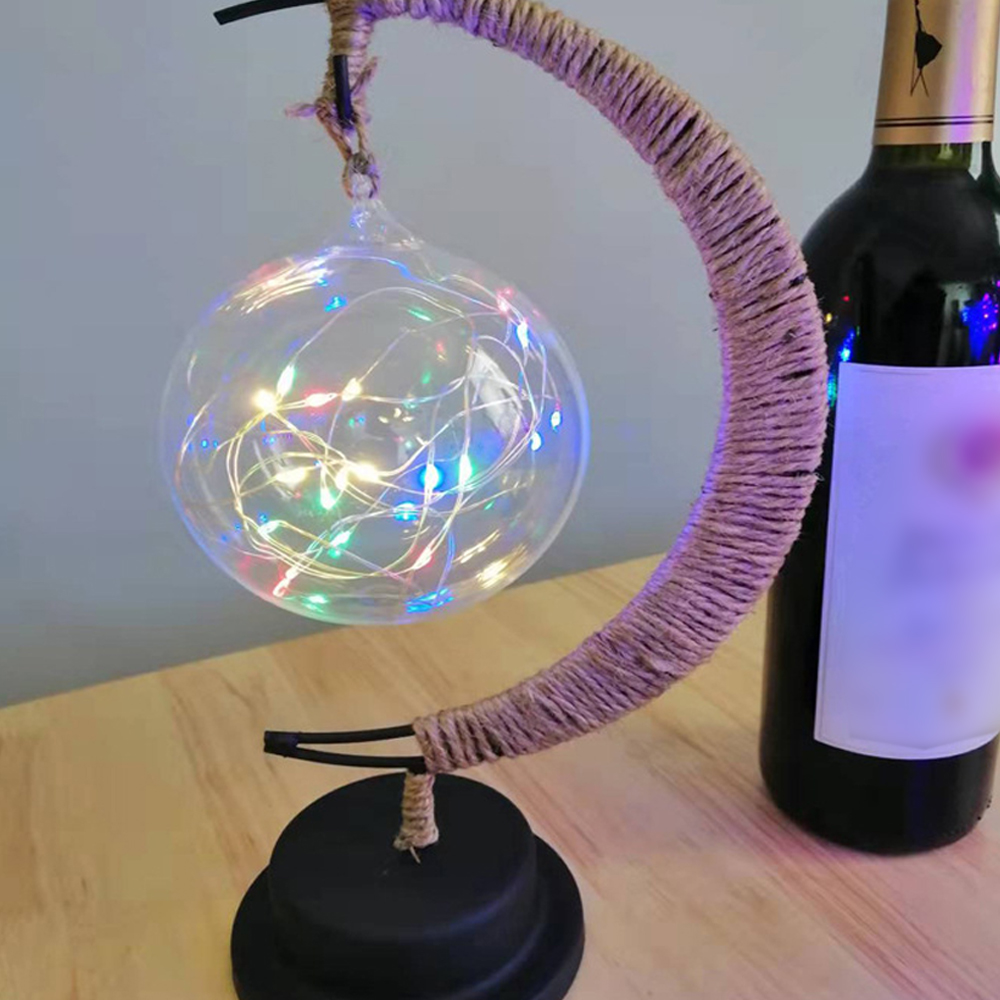 Eine klare Glaskugel mit bunten Lichtern hängt an einem mit Jute umwickelten Metallrahmen neben einer Weinflasche auf einem Holztisch.
