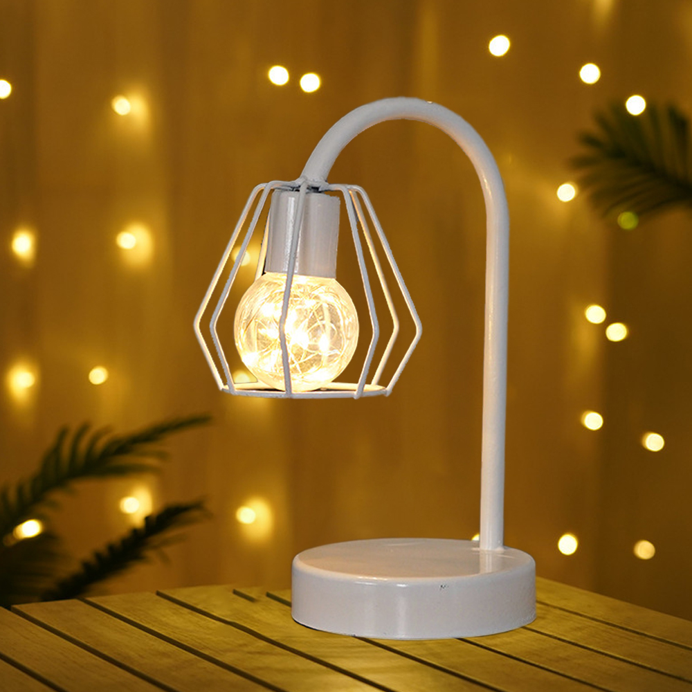 Weiße Lampe mit geometrischem Käfig um die Glühbirne. Sie steht auf einem Holztisch mit Hintergrundlichtern und Pflanzen.