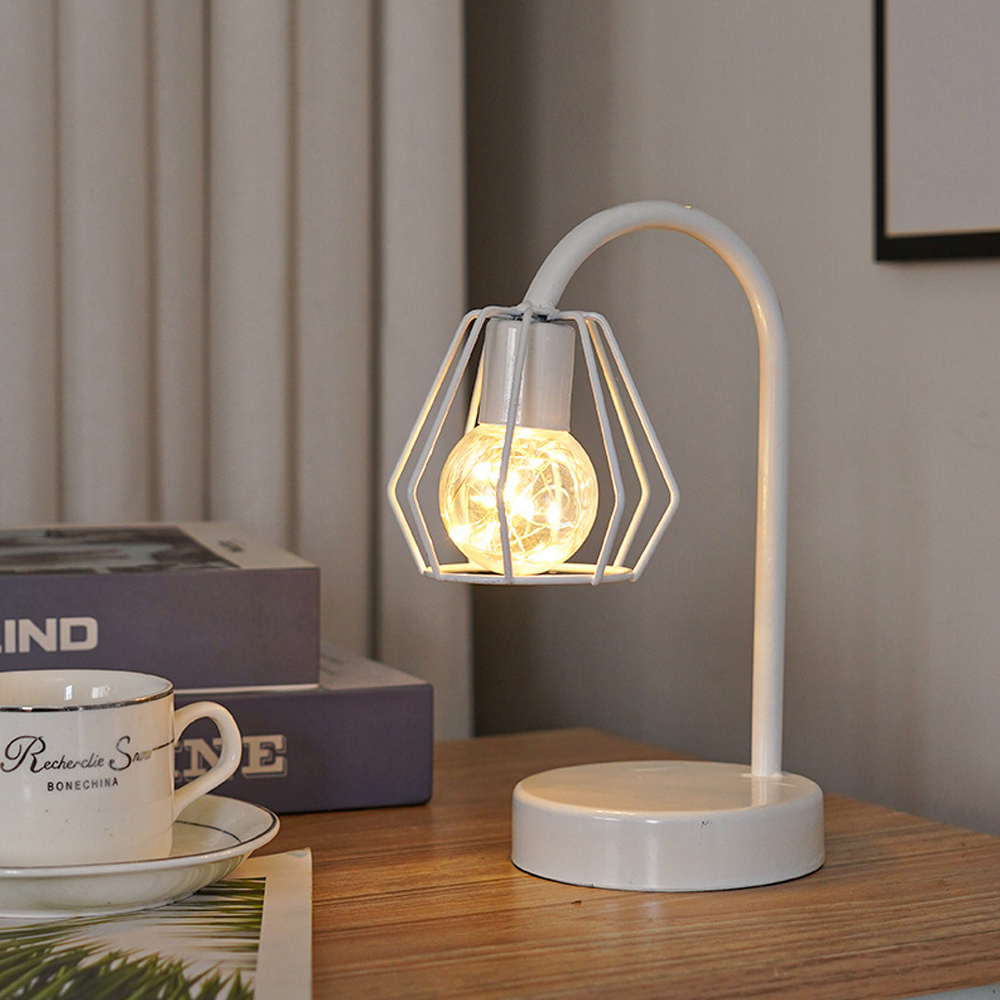 Eine weiße Schreibtischlampe mit geometrischem Schirm. Im Inneren befindet sich eine leuchtende Glühbirne. Auf dem Tisch stehen eine Teetasse und Bücher.