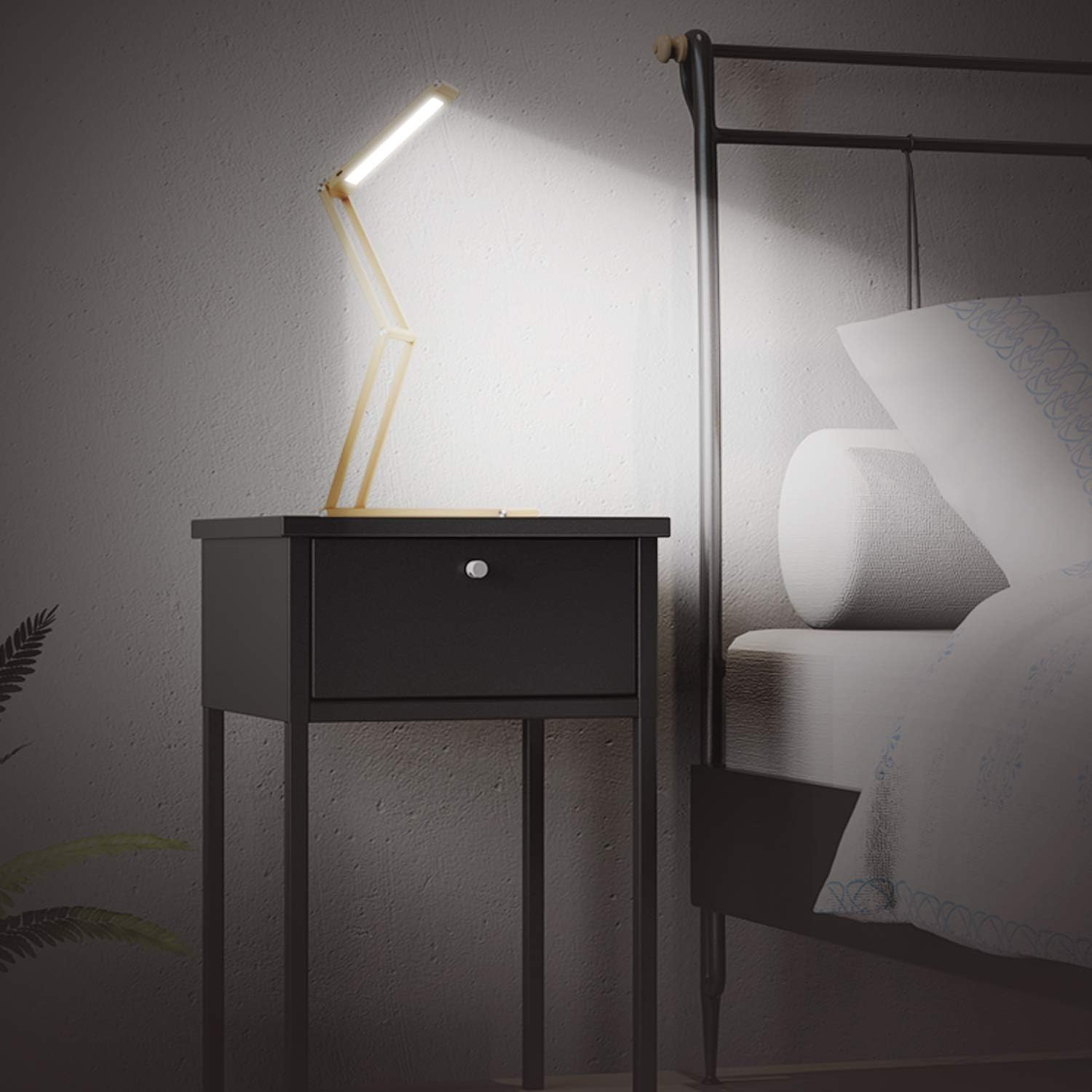 Eine beleuchtete Holzschreibtischlampe auf einem schwarzen Nachttisch. Im Hintergrund ein Bettgestell mit Kissen und Decke.
