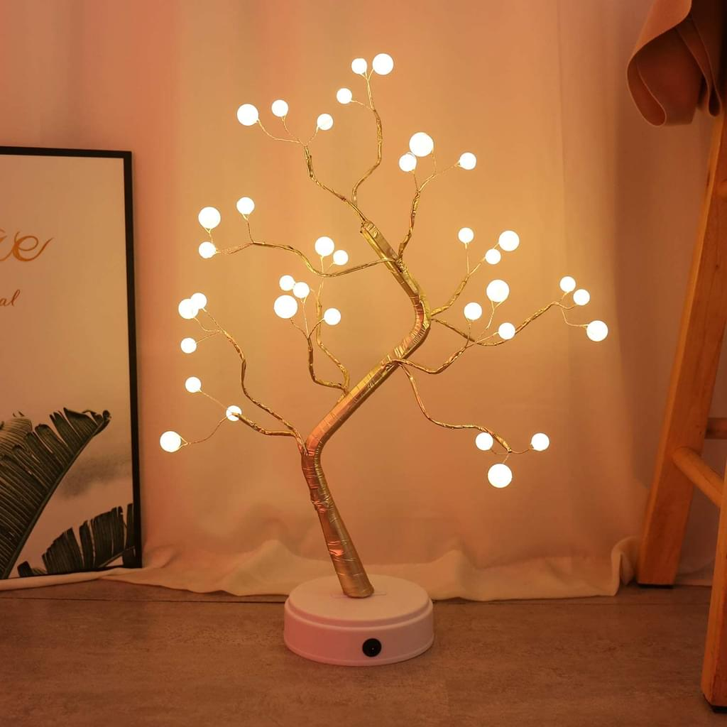 Eine dekorative Baumlampe mit weißen Lichtern. Sie ist goldfarben und steht auf einem weißen Sockel. Im Hintergrund ein gerahmtes Bild und eine Leiter.