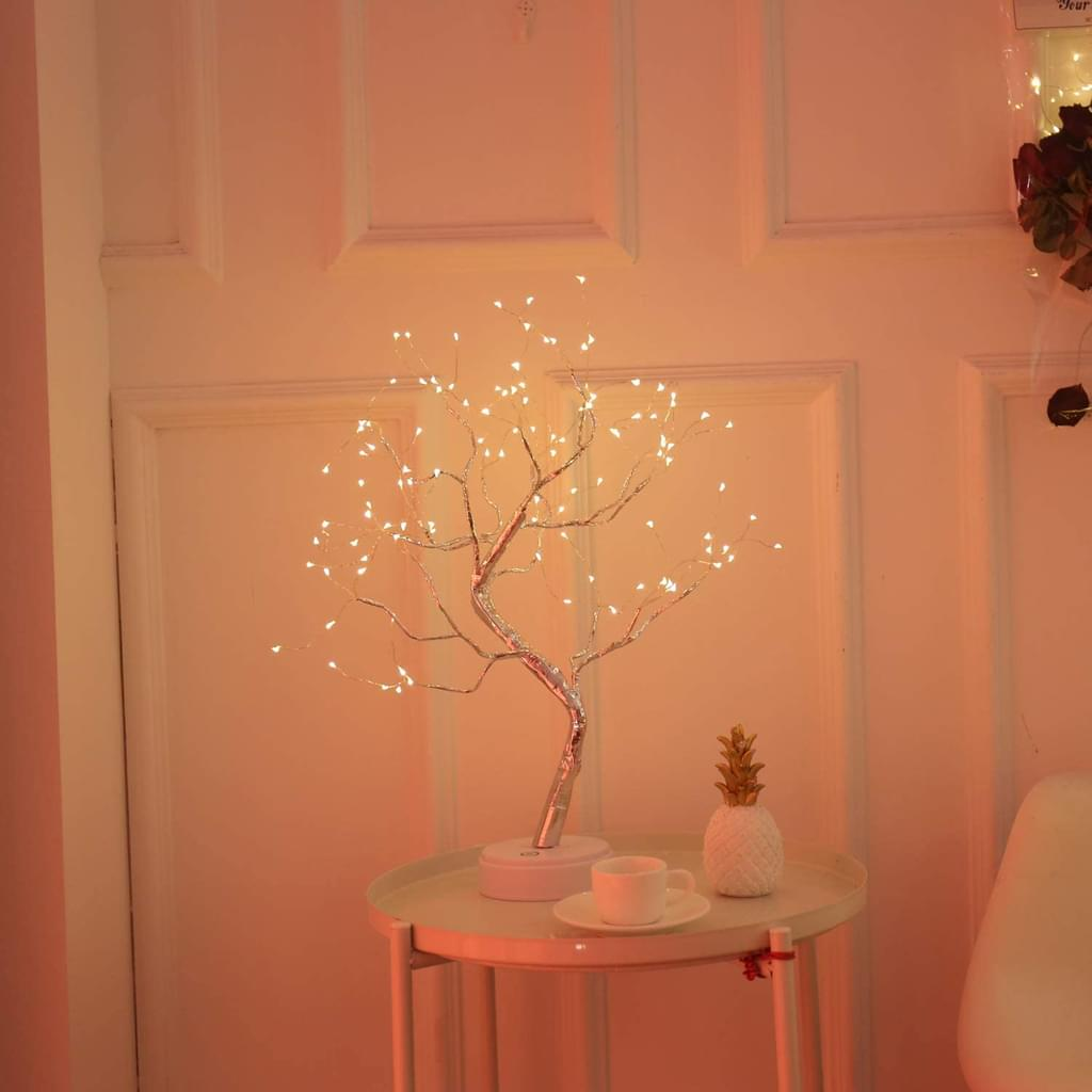 Eine beleuchtete Baumlampe steht auf einem kleinen Tisch. Eine weiße Tasse und eine Ananas-Ornamente stehen auf dem Tisch. Der Hintergrund ist eine Tür.