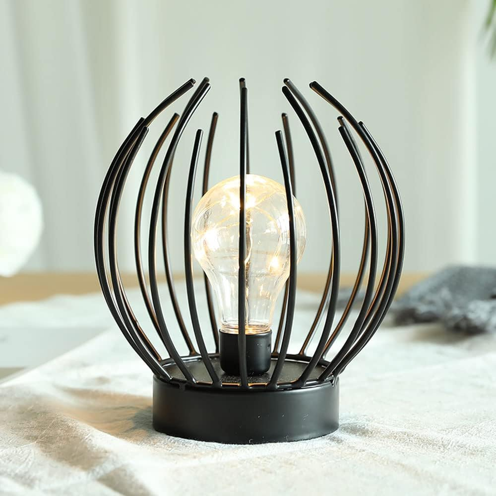 Eine dekorative Lampe mit einer Glühbirne in einem schwarzen Metallkäfig. Sie steht auf einer weißen Oberfläche. Der Hintergrund ist unscharf.