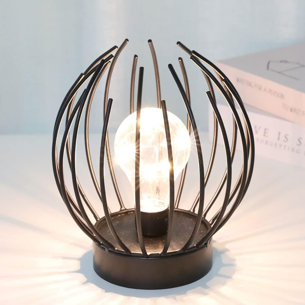 Eine schwarze Metallkäfiglampe mit einer glühenden Glühbirne im Inneren. Die Lampe steht auf einer weißen Oberfläche. Bücher sind im Hintergrund.