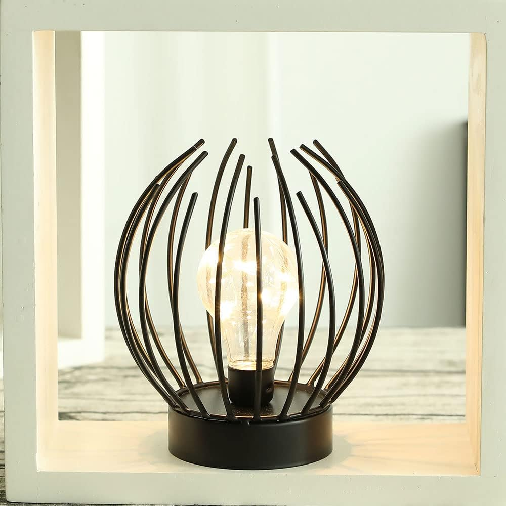 Eine dekorative Lampe mit einer Glühbirne, die in einem schwarzen Metallkäfig eingeschlossen ist. Die Lampe steht auf einem schwarzen Sockel, in einem weißen Rahmen.