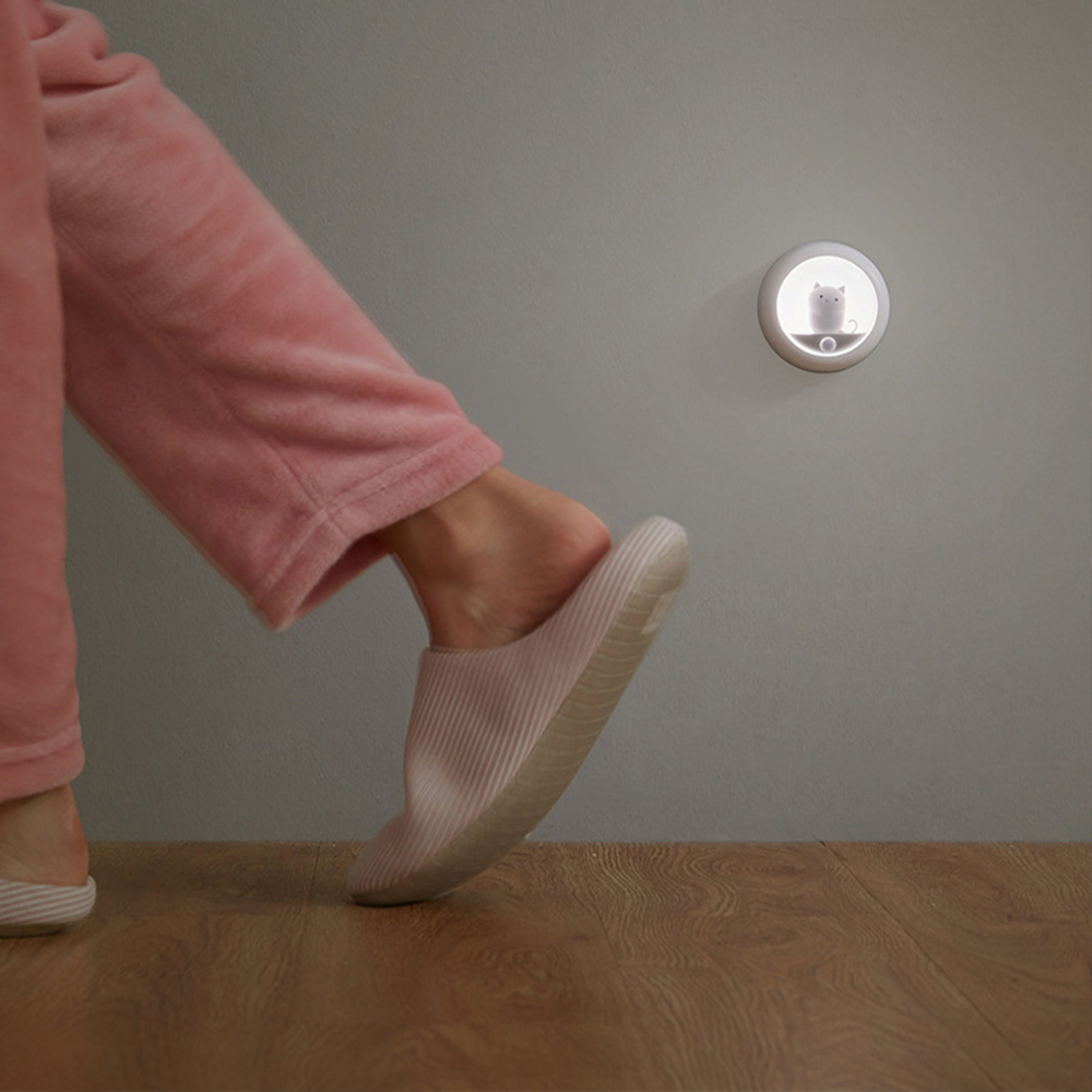 Eine Person in rosa Pyjama und Hausschuhen geht. Eine Wandleuchte mit Katzendesign ist an der Wand. Holzfußboden.