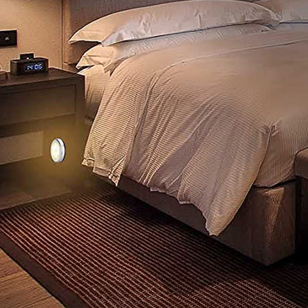 Schlafzimmer mit Bett, Nachttisch mit Uhr und Teppich. Ein kleines Licht scheint unter dem Tisch.