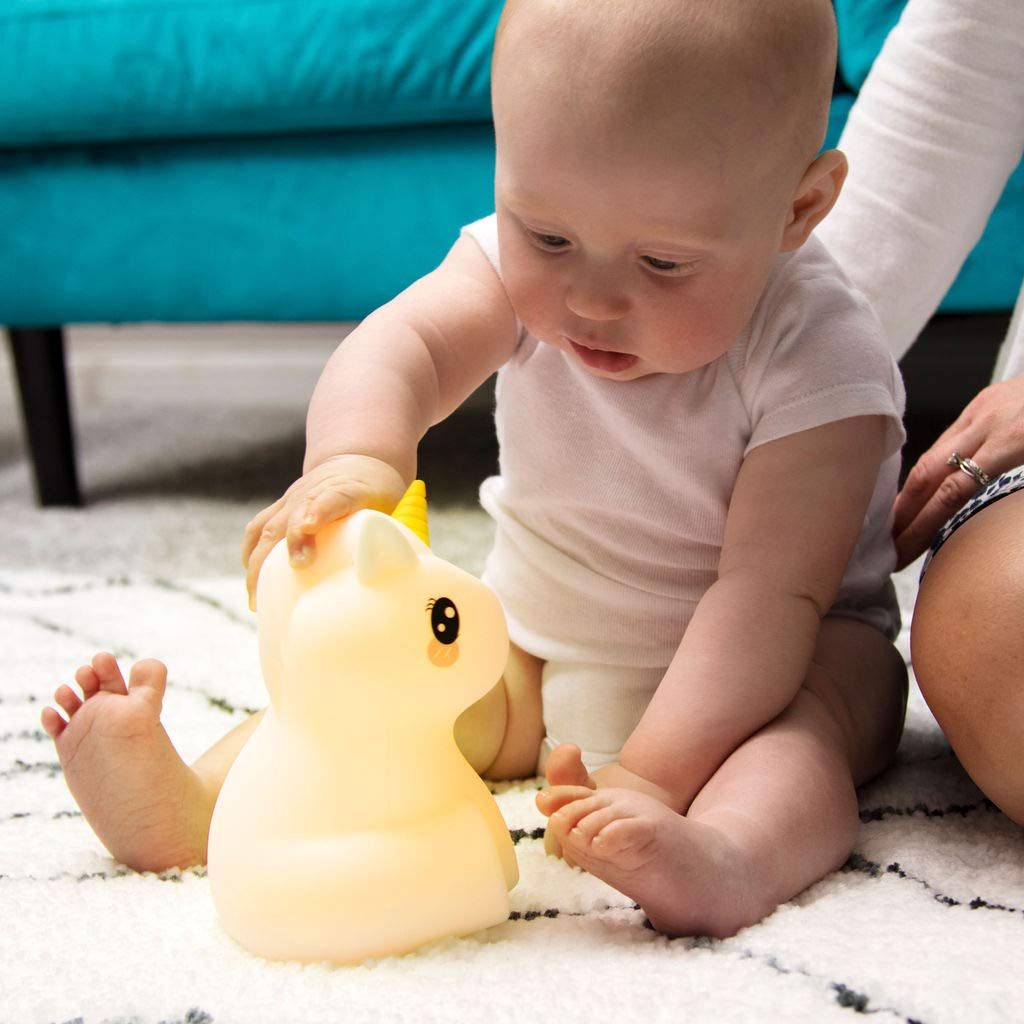 Baby im weißen Strampler berührt eine leuchtende Einhornlampe auf einem Teppich. Eine blaue Couch und die Beine einer Person sind im Hintergrund.