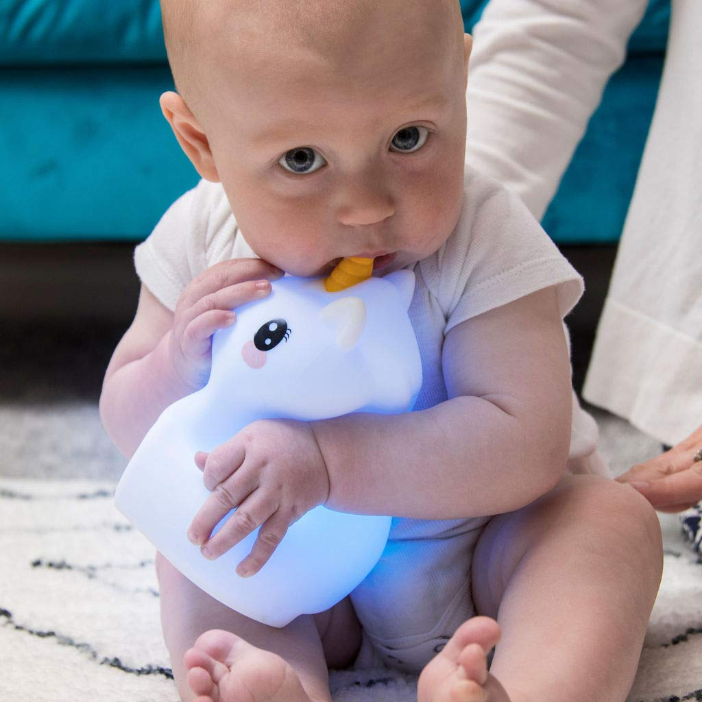Ein Baby in einem weißen Strampler sitzt auf einem Teppich und hält ein leuchtendes weißes Einhorn-Spielzeug mit gelbem Horn und beißt darauf.