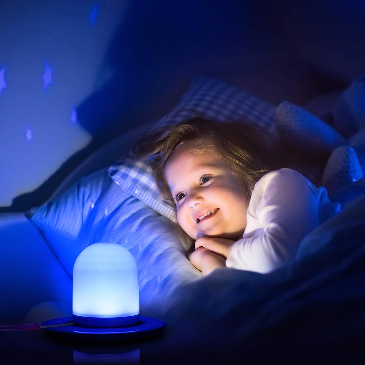 Ein Kind lächelt im Bett, beleuchtet von einer blauen Lampe. Sternprojektionen an der Wand. Das Kind ist von Kissen und einem Teddybär umgeben.