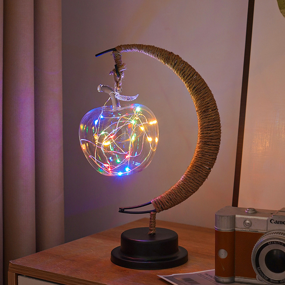 Eine glasapfelförmige Lampe mit bunten Lichtern hängt an einem halbmondförmigen Ständer. Eine Kamera steht auf einem Holztisch.