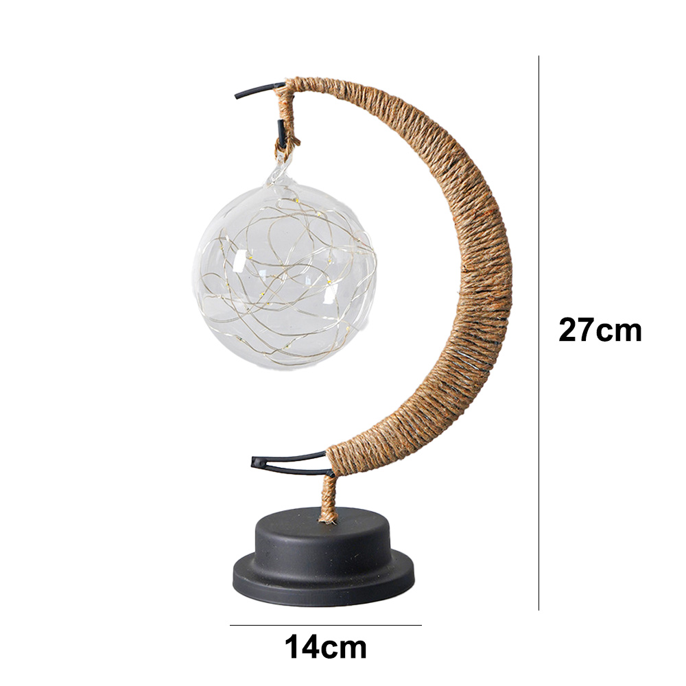 Eine dekorative Lampe. Eine Glaskugel mit Lichterketten hängt an einem gewebten Halbmond. Sie hat einen schwarzen Sockel. Maße sind angegeben.