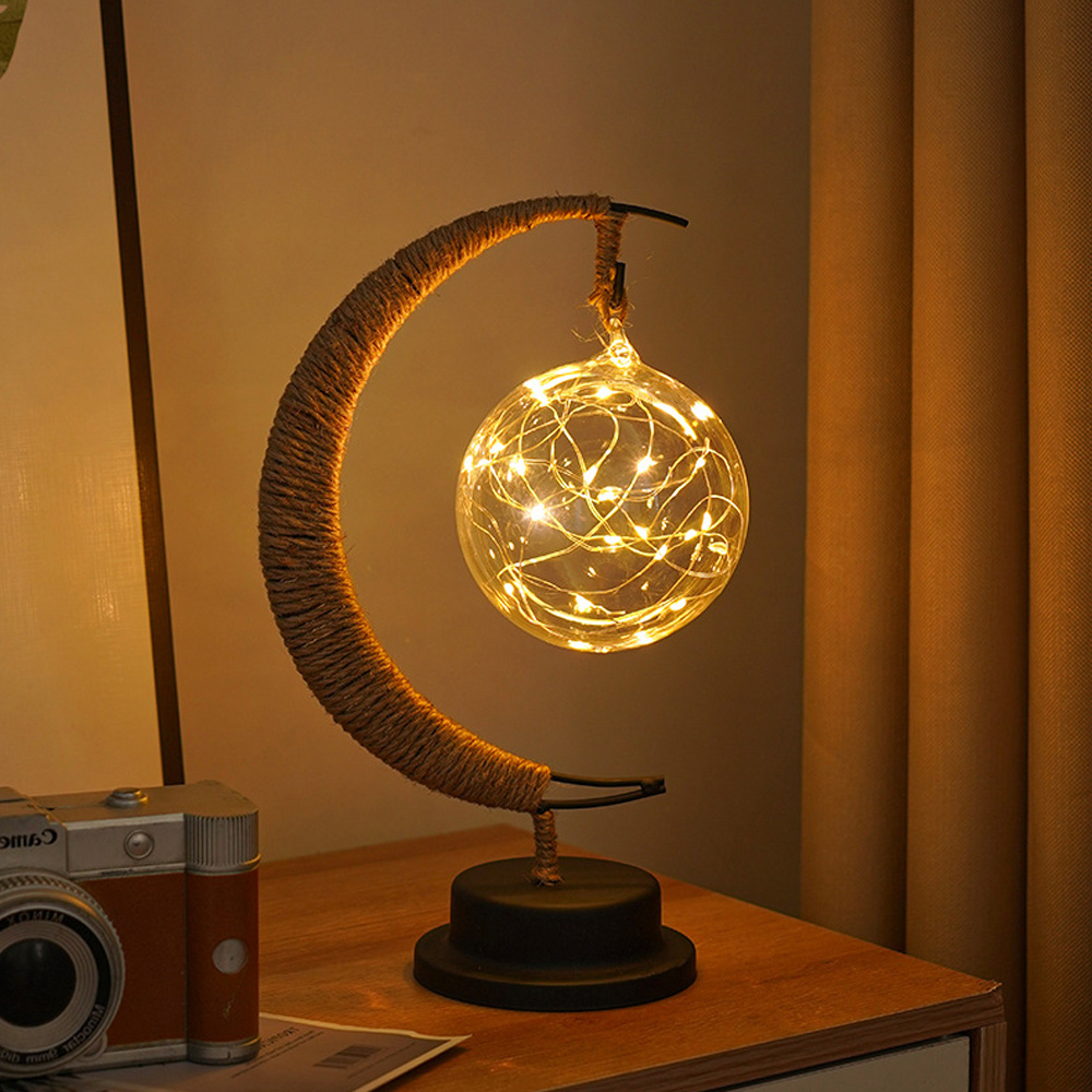 Eine dekorative Lampe mit einem halbmondförmigen Seilrahmen, der eine Glaskugel mit Lichterketten hält. Auf einer Holzoberfläche befindet sich eine Kamera.