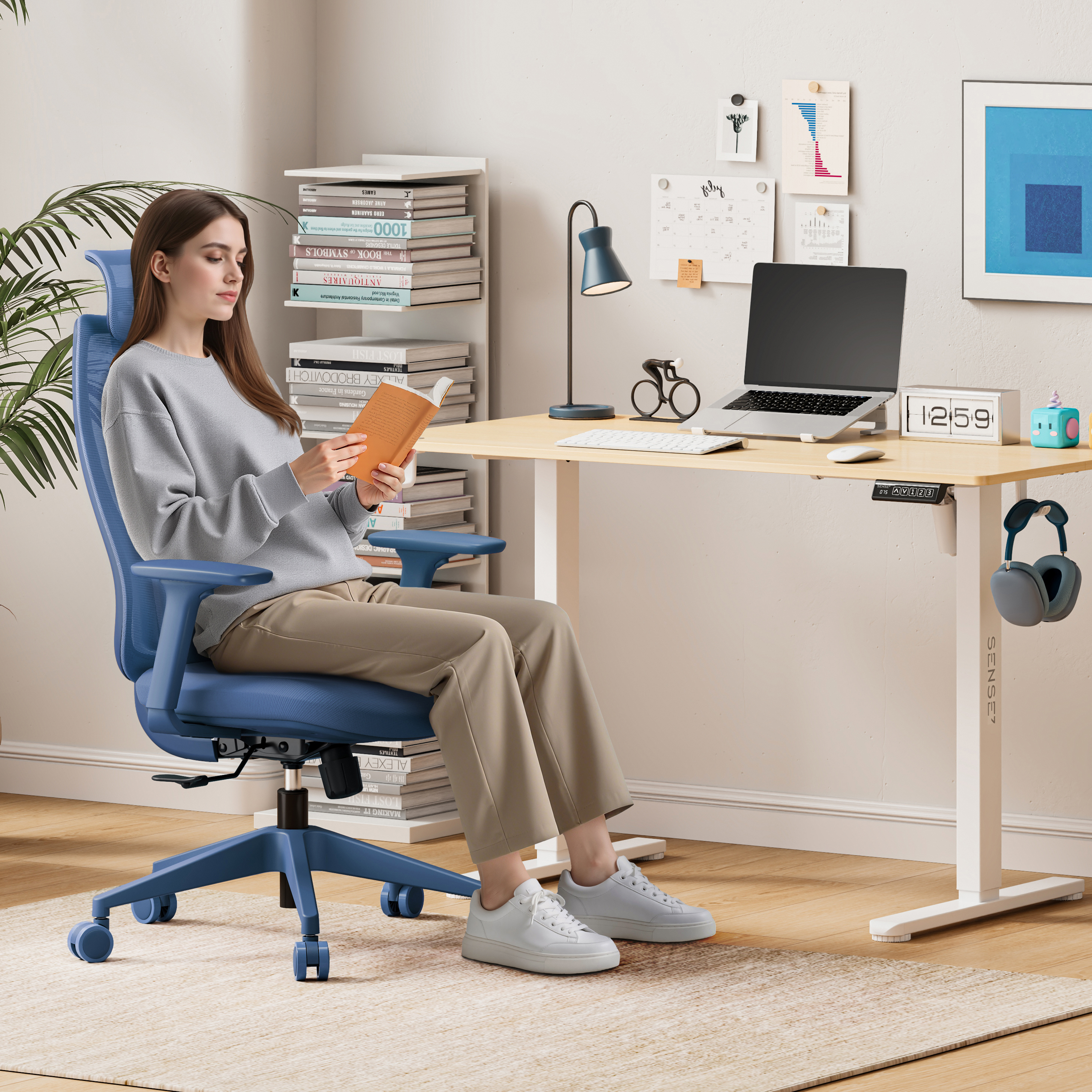 Una donna legge un libro su una sedia blu, a una scrivania con un laptop e una lampada. Ci sono librerie dietro di lei.