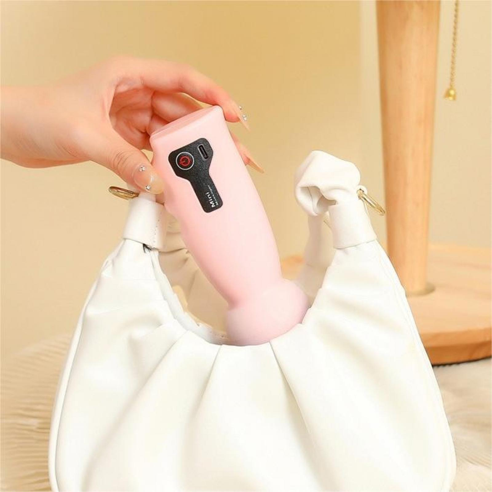 Eine Hand hält ein rosa Gerät und legt es in eine weiße Handtasche. Ein Holzständer ist im Hintergrund.
