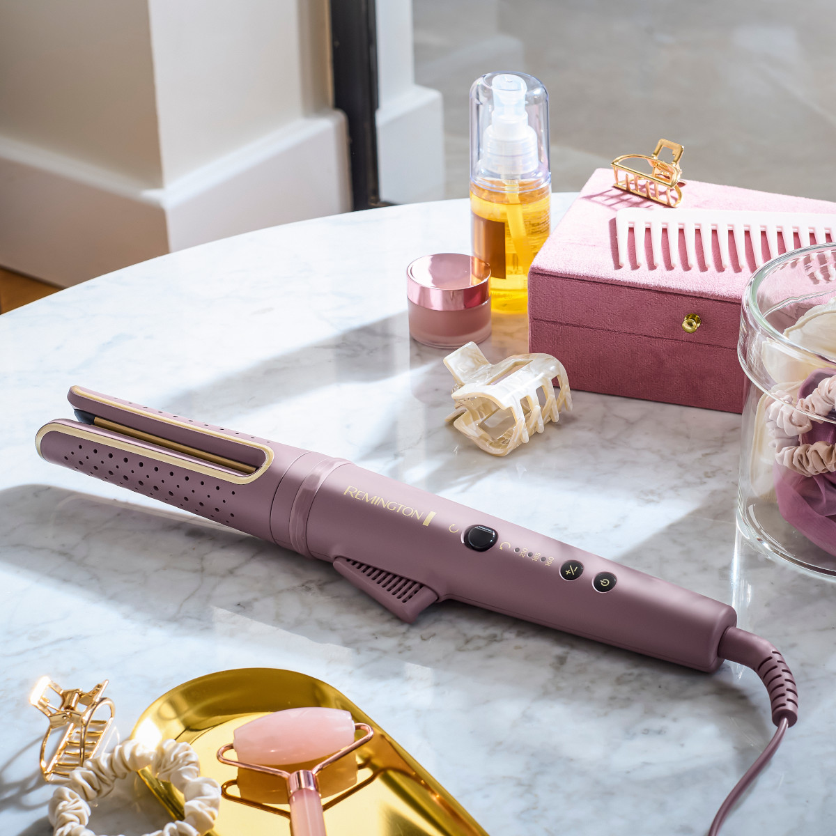 Una piastra, un rullo di quarzo rosa e accessori per capelli su un tavolo di marmo. Presenti anche un portagioie rosa e olio per capelli.