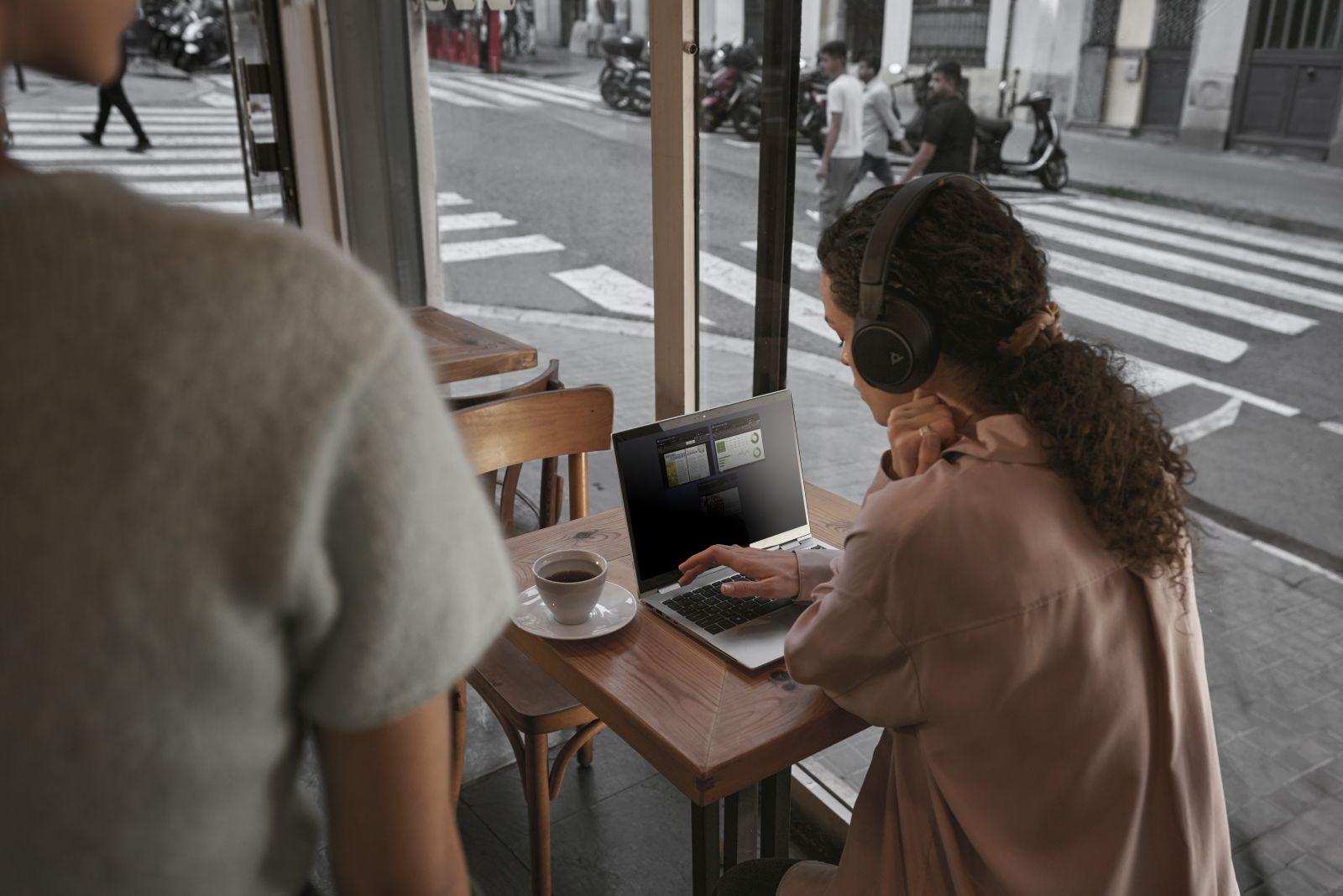 Donna con cuffie usa il laptop al tavolo del bar. Tazza di caffè sul tavolo. Scena di strada visibile dalla finestra.