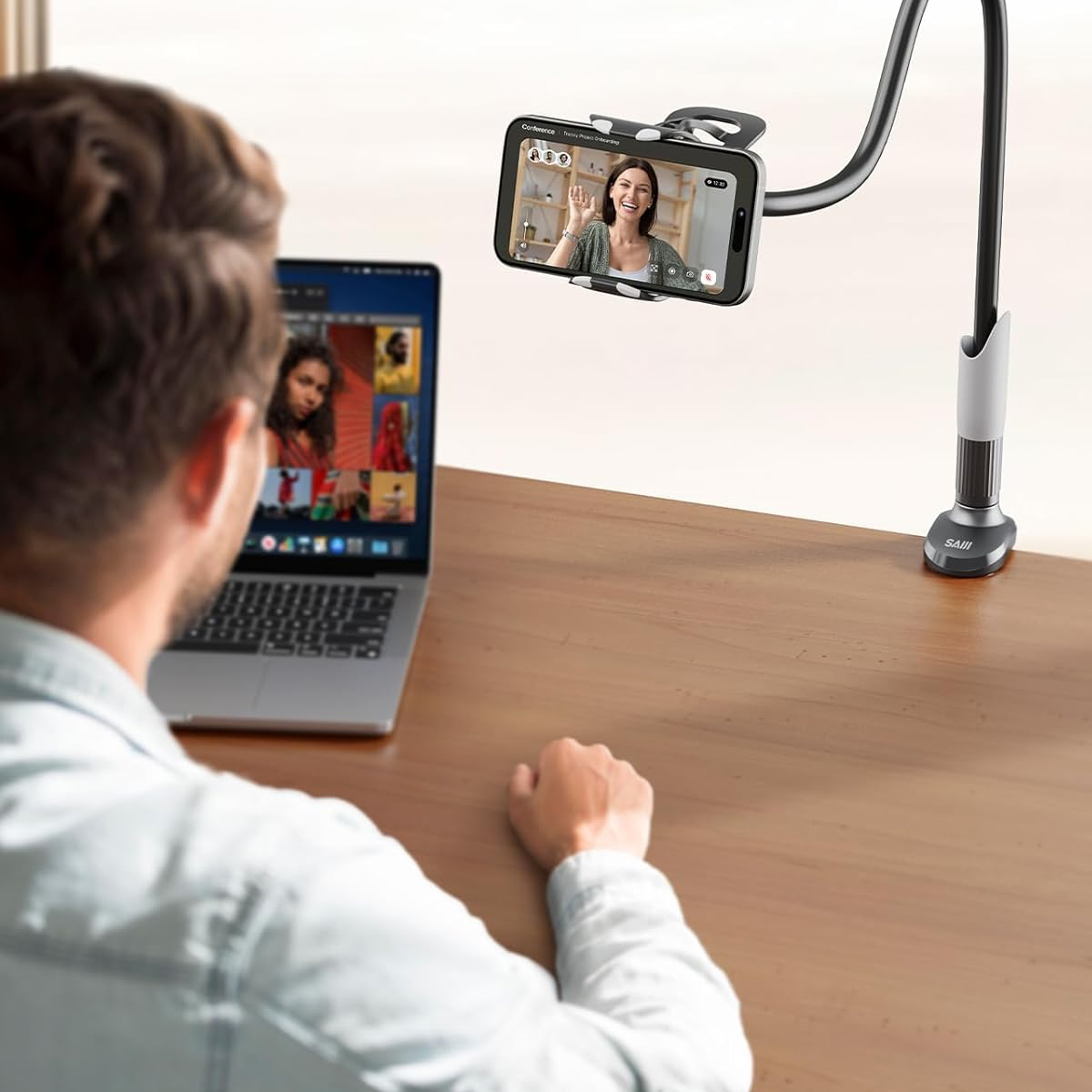 Mann bei Videoanruf mit Frau auf Telefon. Telefon an flexiblem Ständer, Laptop zeigt weitere Teilnehmer auf dem Tisch.