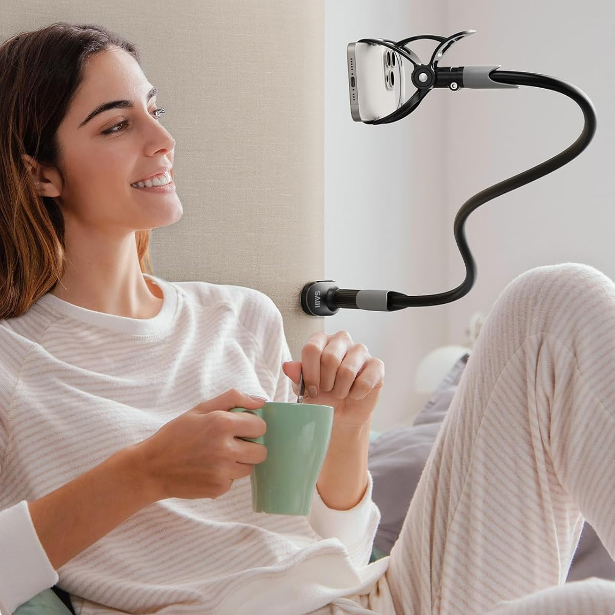 Frau im Pyjama hält eine Tasse und lächelt. Ein Telefon wird von einem flexiblen Ständer gehalten. Die Wand ist beige.