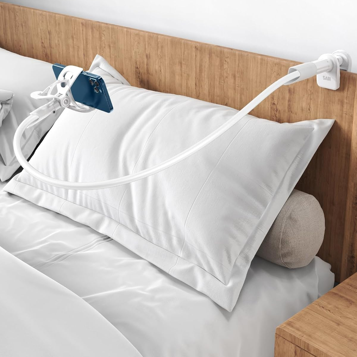 Ein Telefon wird von einem weißen, flexiblen Arm gehalten, der an einem Kopfteil befestigt ist. Das Telefon befindet sich über einem weißen Kissen auf einem Bett.