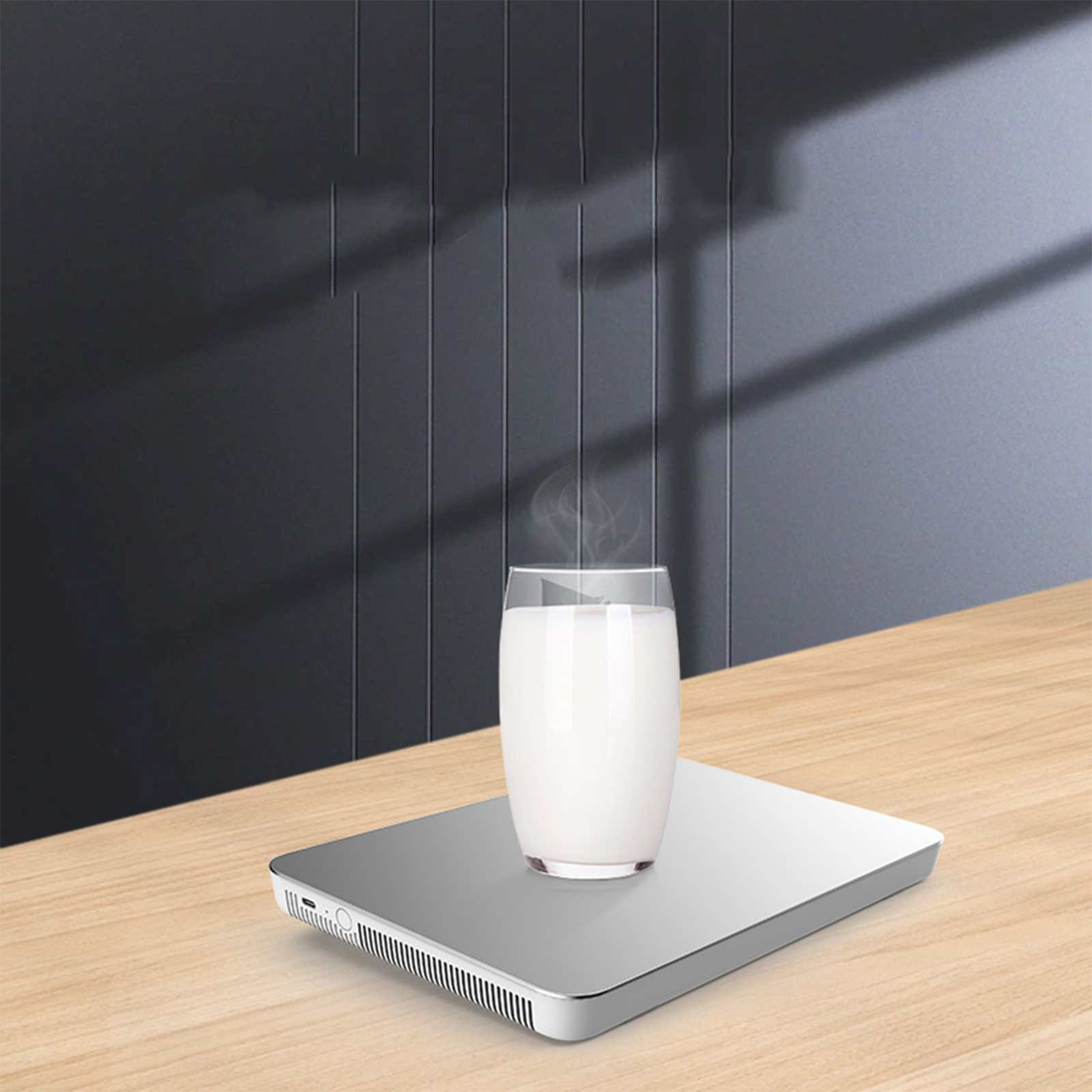 Ein Glas Milch auf einem silbernen Wärmer. Dampf steigt von der Milch auf. Der Hintergrund ist eine dunkelgraue Wand und ein Holztisch.