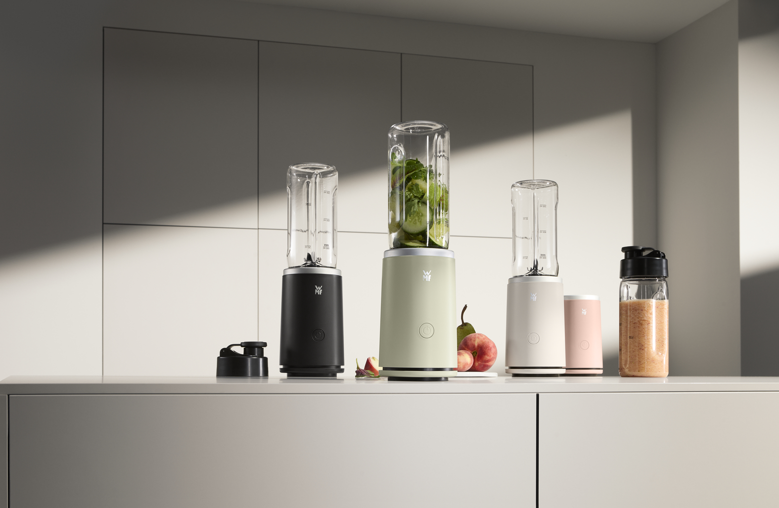 Mehrere Mixer in verschiedenen Farben mit Gläsern und Obst. Vordergrund: schwarze, hellgrüne und weiße Mixer. Hintergrund: hellgraue Wand.