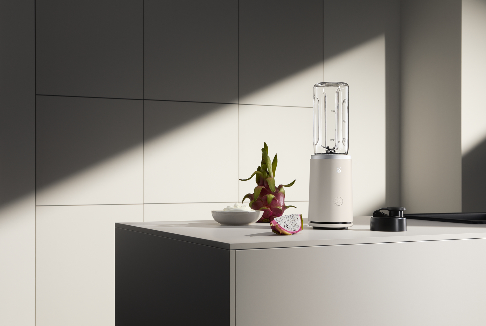 Eine Küchenszene mit einem Mixer, Drachenfrucht und einer kleinen Schüssel mit weißer Creme auf einer weißen Arbeitsplatte.