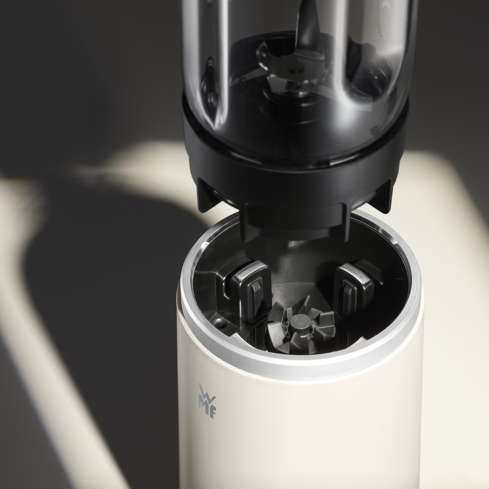 Ein Mixer, zerlegt. Der obere Teil mit Klingen befindet sich über der Basis. Beigefarbene Basis, silberner Rand, schwarze Klingen und Oberteil.