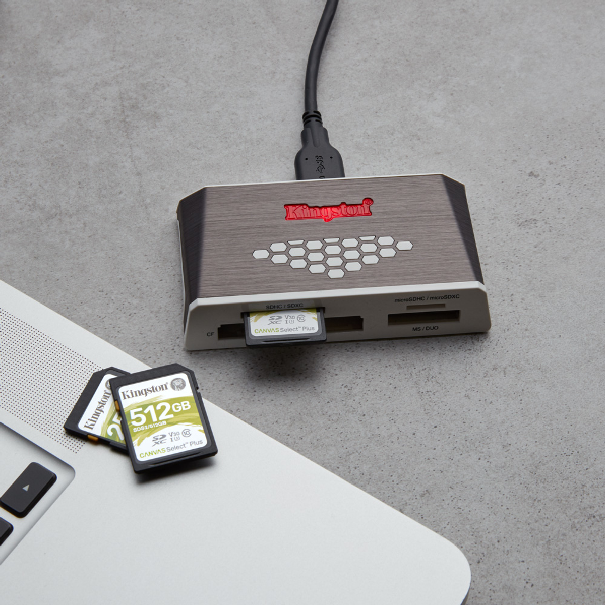Un lector de tarjetas Kingston con cable USB conectado. Tarjetas SD y un portátil sobre una superficie gris.
