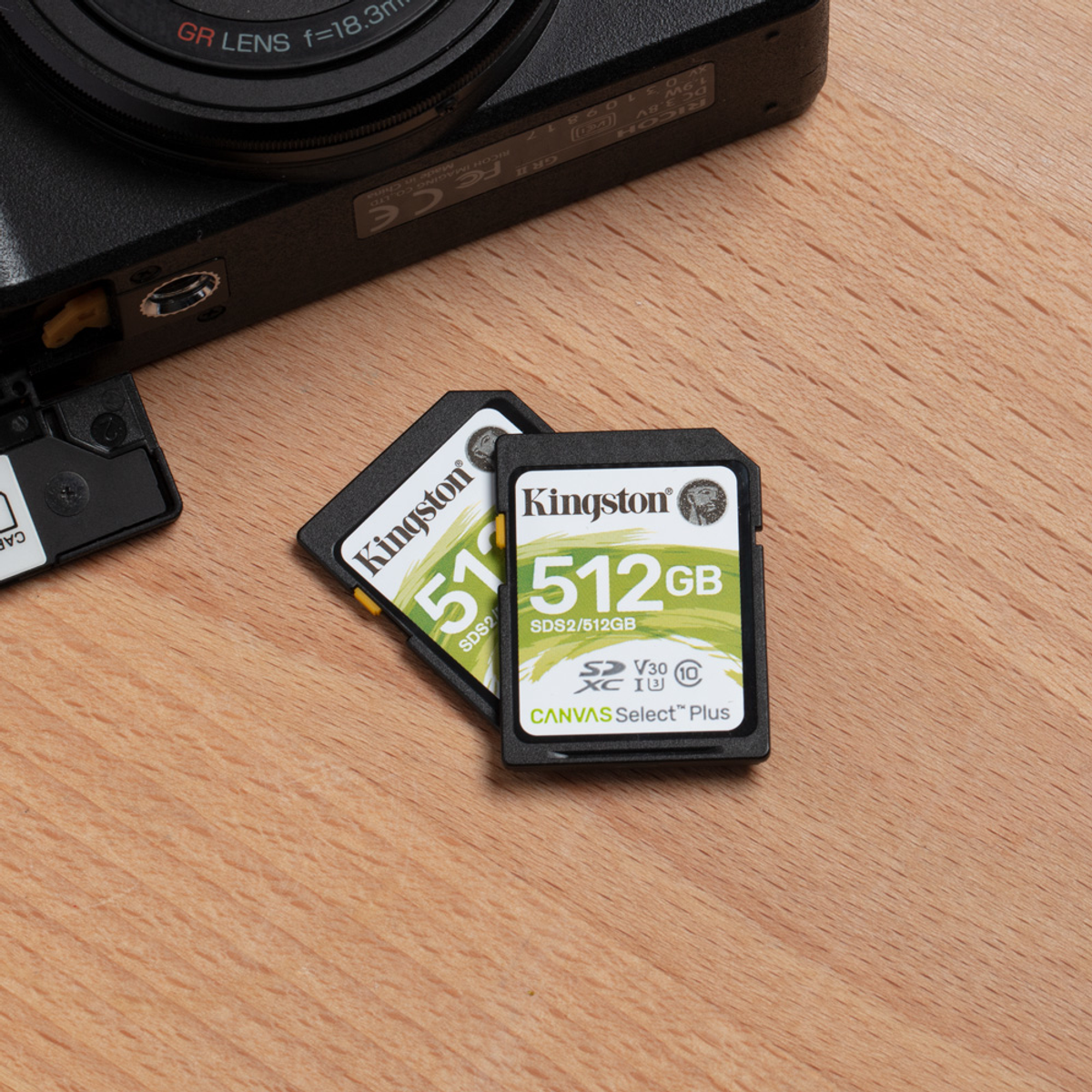 Dos tarjetas SD Kingston de 512 GB sobre una superficie de madera, cerca de una lente de cámara y una ranura para tarjetas.