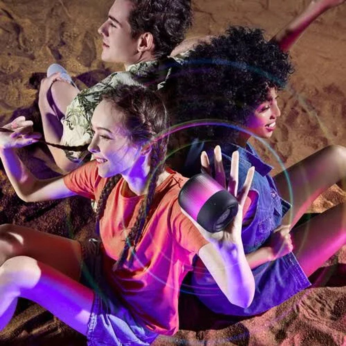 Trzy osoby siedzą na piasku, trzymają głośnik. Otaczają je tęczowe światła. Jedna dziewczyna w pomarańczowym, inne w dżinsie i kwiatach.