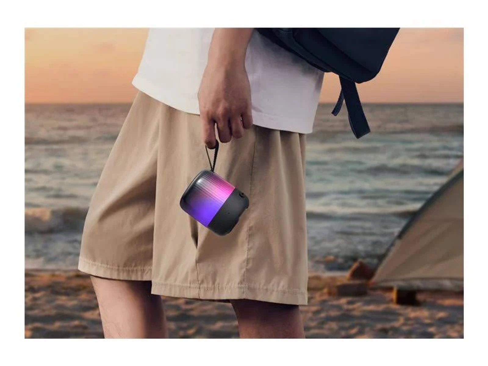 Osoba trzyma głośnik z kolorowymi światłami. Ma na sobie szorty, białą koszulę i plecak. W tle plaża i namiot.