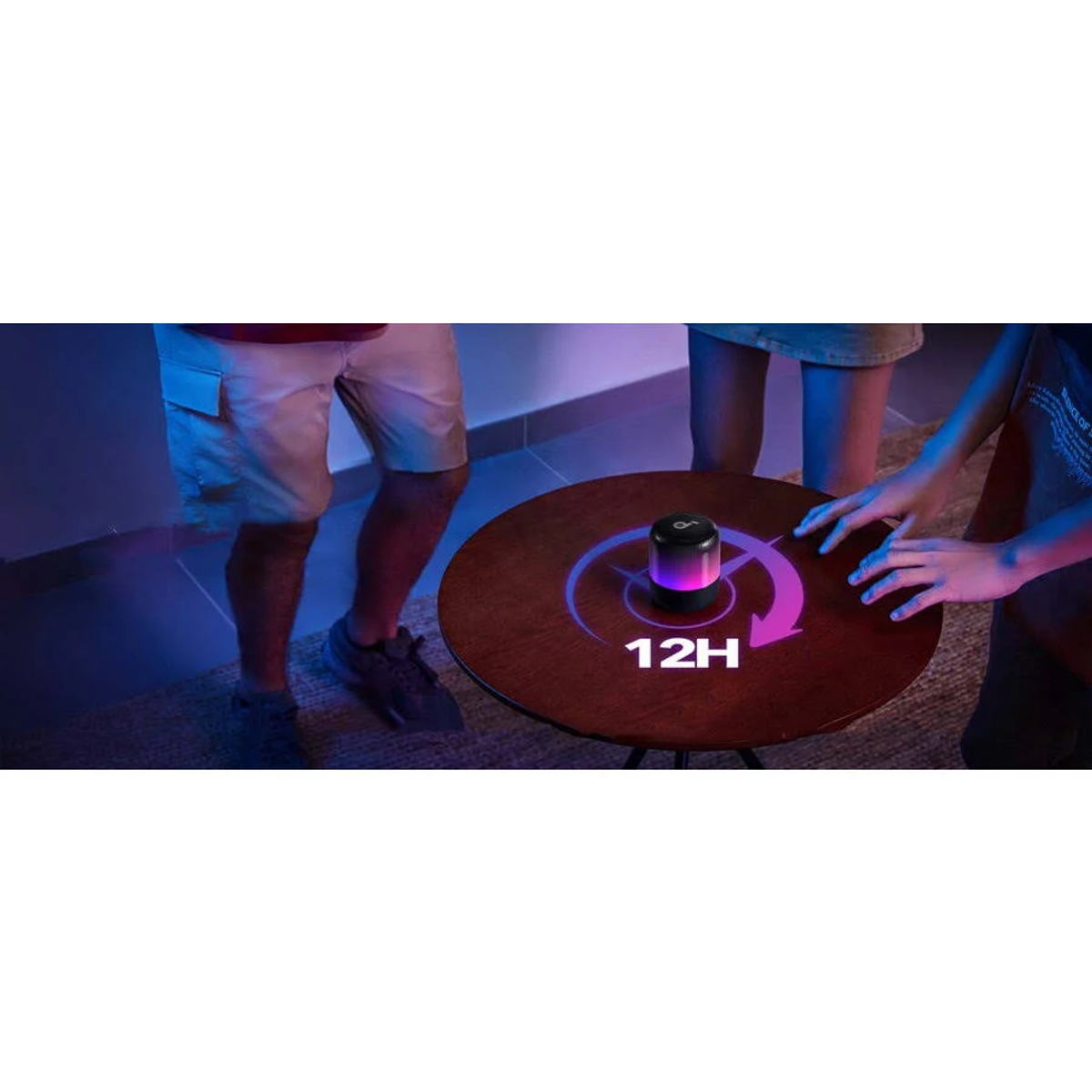 Czarny głośnik na okrągłym stole z ludźmi wokół. Na stole napisano 12H. Widoczne jest fioletowe i różowe światło.