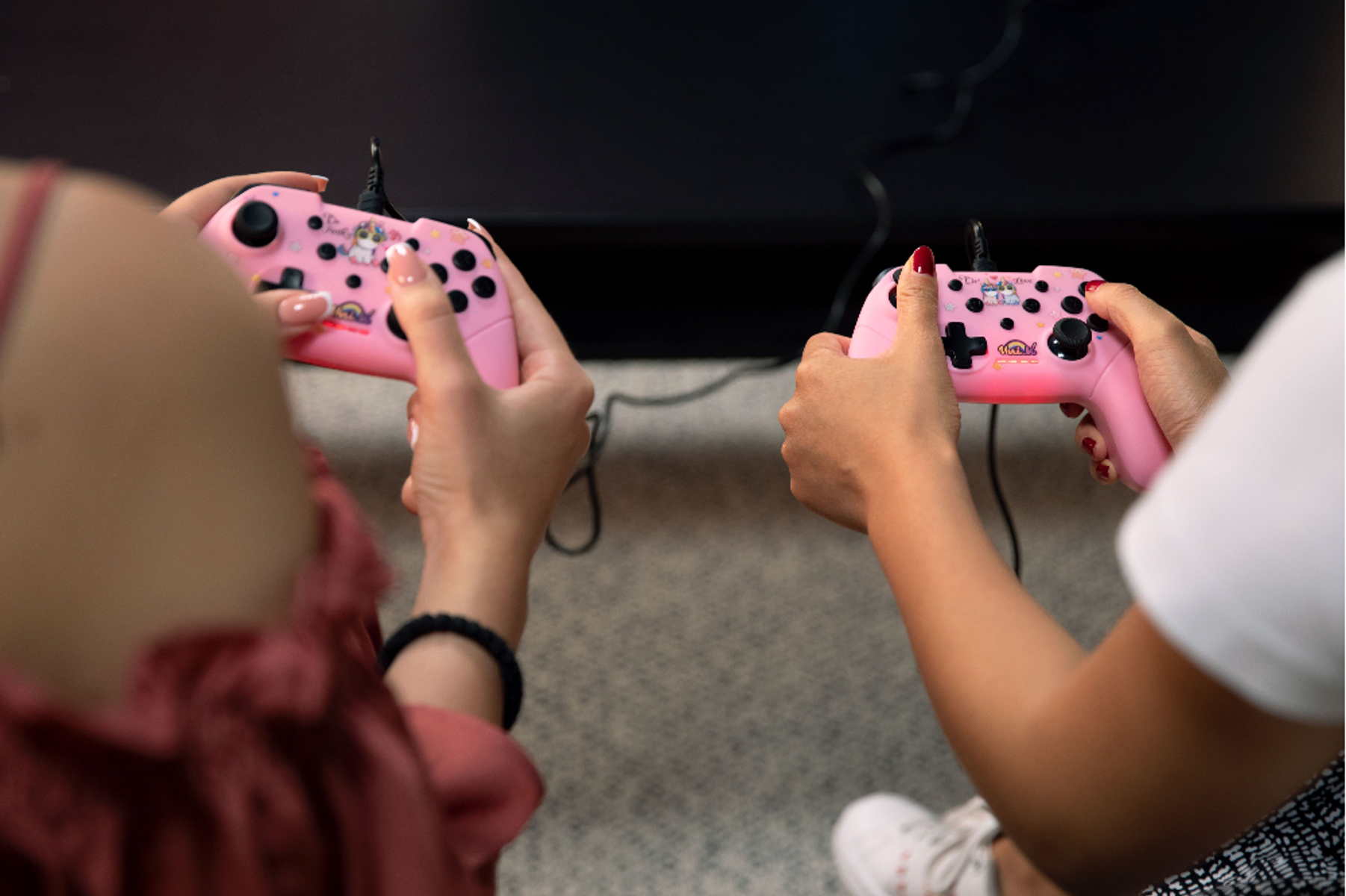 Dwie osoby trzymają różowe kontrolery do gier z czarnymi przyciskami, grając. Dłonie i kontrolery są ostre.