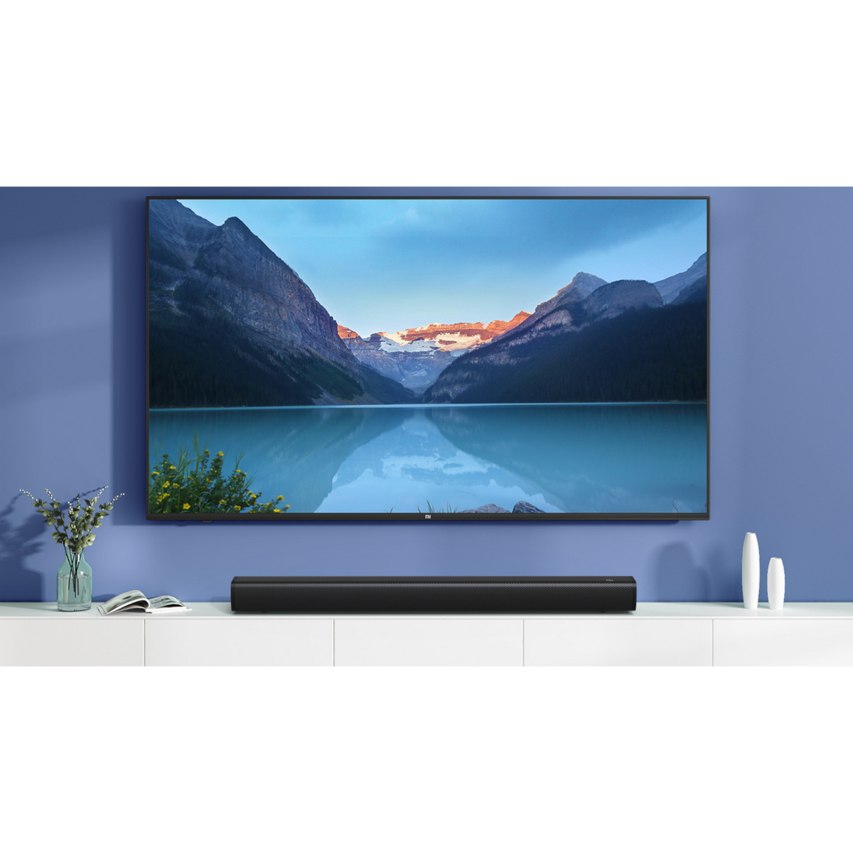 Telewizor wyświetla scenę jeziora. Poniżej soundbar, na białej szafce. Wazy i kwiaty stoją na szafce.