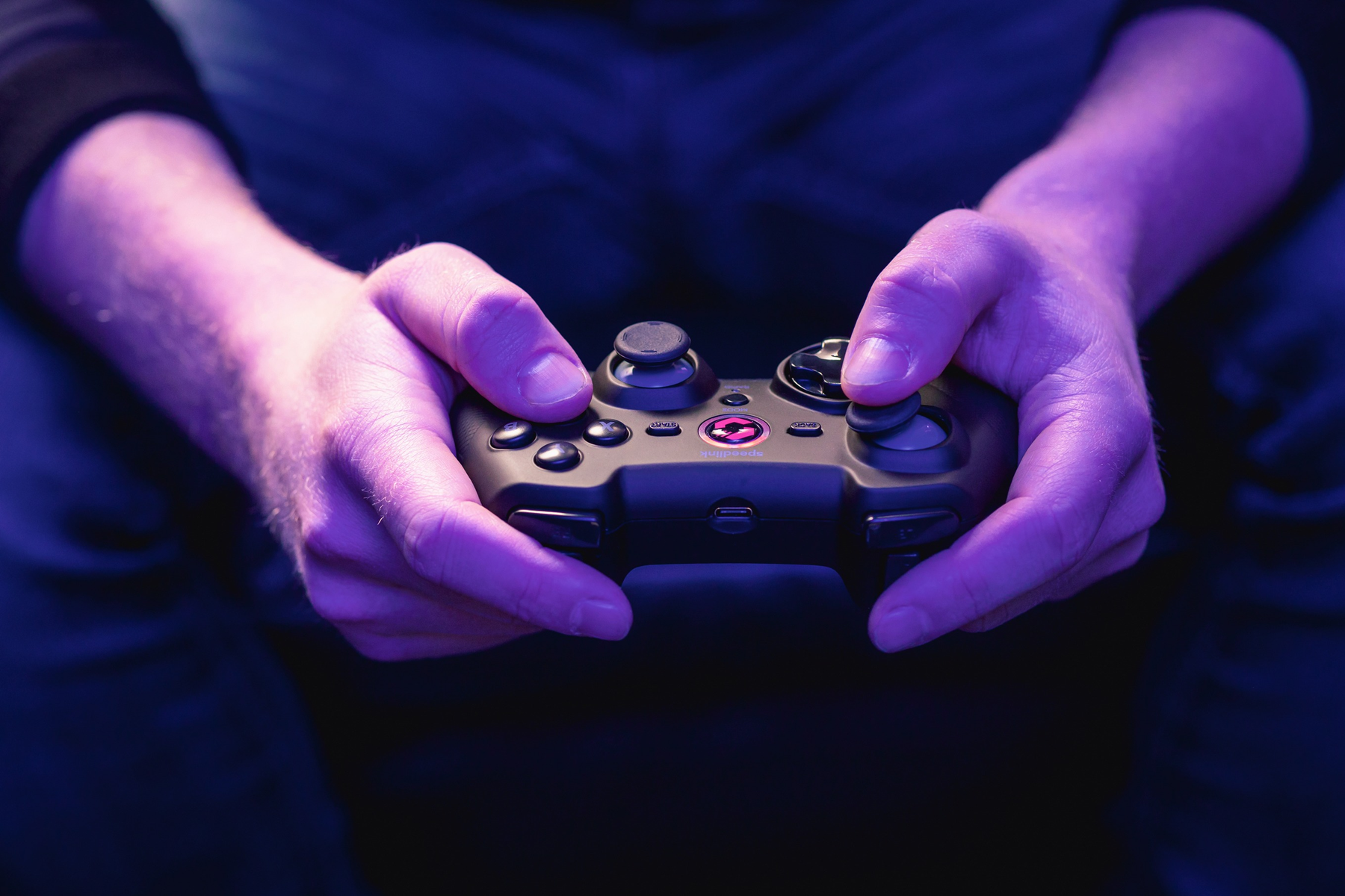 Dłonie trzymają czarny kontroler do gier z różowymi akcentami. Tło jest rozmyte, ciemnoniebieskie.