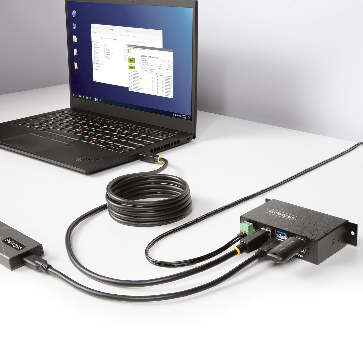 Un portátil conectado a un concentrador negro mediante cables. La pantalla del portátil muestra un explorador de archivos y una ventana de información del sistema.