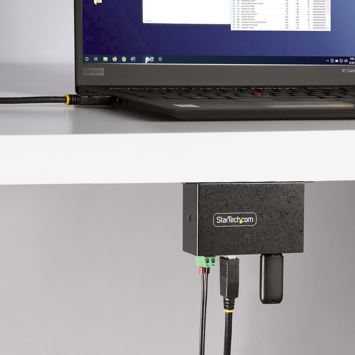 Una laptop Lenovo conectada a una caja negra StarTech.com con cables. La pantalla muestra datos. La caja está montada debajo de una superficie blanca.