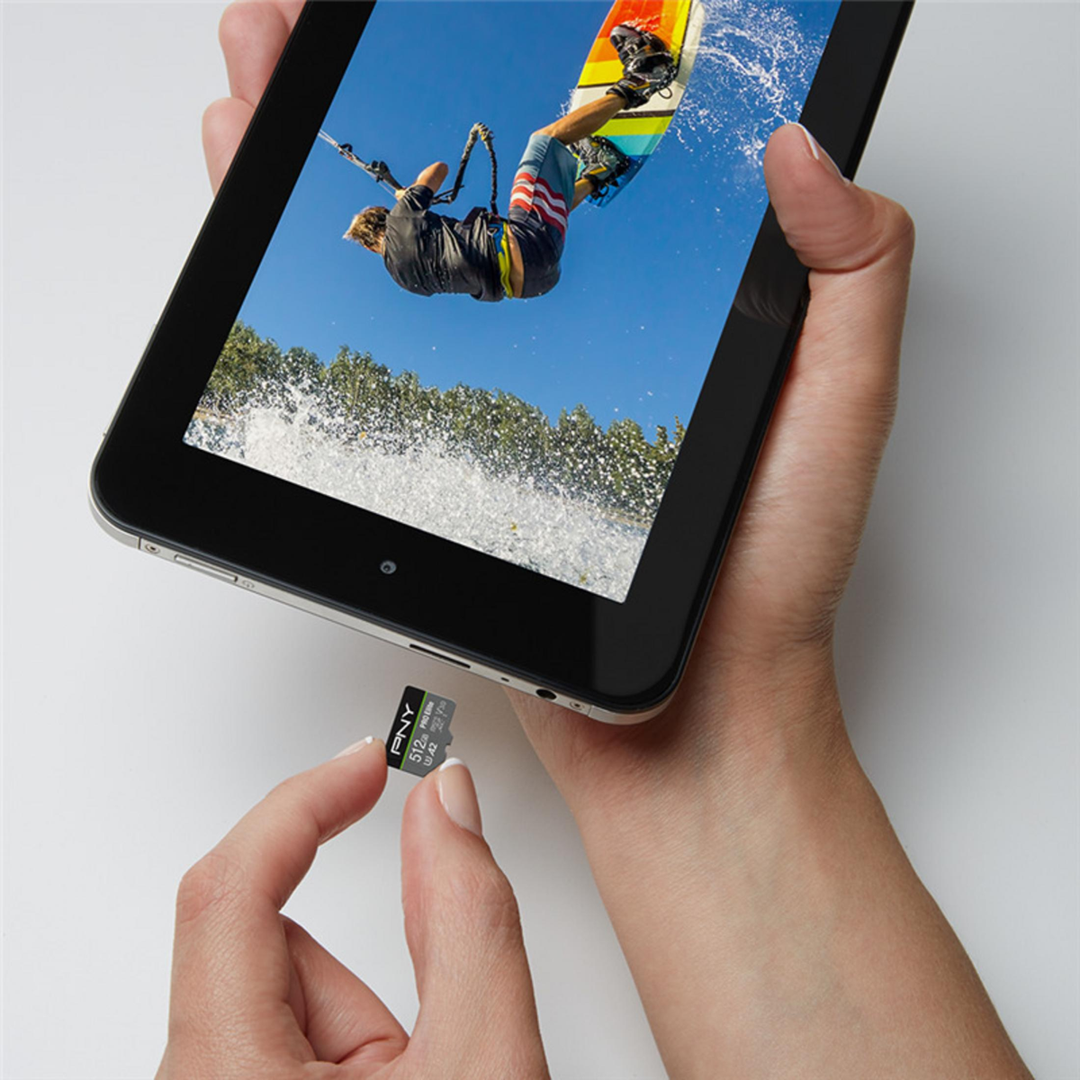 Hände halten ein Tablet mit einem Kitesurfer. Eine microSD-Karte wird in der Nähe des Tablets gehalten. Weißer Hintergrund.