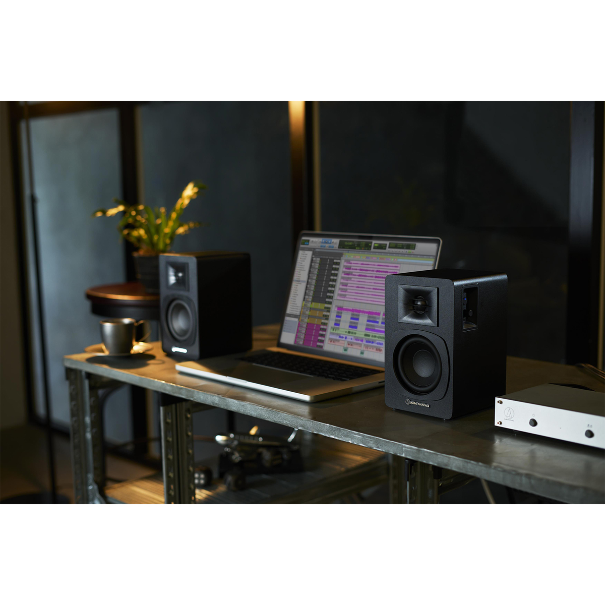 Ustawienia studia z dwoma czarnymi głośnikami, laptopem z oprogramowaniem do edycji audio i filiżanką na metalowym stole.